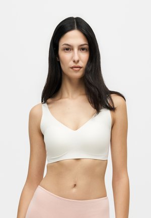 Femme aux longs cheveux foncés portant un crop top blanc sans manches et un pantalon rose clair taille basse sur fond blanc uni.