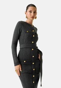 Vestito a maglia grigio strutturato con maniche lunghe, chiusura frontale con bottoni, due tasche sul petto e cintura coordinata. Bottoni e dettagli dorati su tutto il capo.