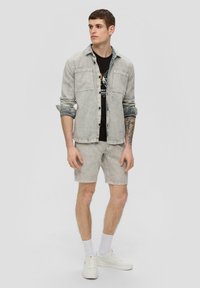 Lichtgrijze denimjack met twee borstzakken, gedragen over een zwart T-shirt. Bijpassende lichtgrijze denimshorts en witte sneakers, met zichtbare tatoeages.