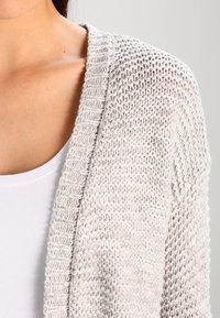 Cardigan lavorato a maglia grigio chiaro con un motivo testurizzato, apertura frontale e dettagli a coste lungo i bordi. Presenta una vestibilità rilassata.