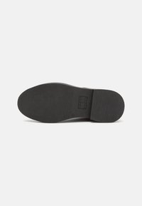 Suela de zapato de goma negra con ranuras horizontales, punta redondeada y tacón plano, con un emblema imprimido en la parte inferior.
