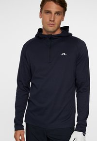 Marinblå långärmad tröja med halv dragkedja och huva. Har en liten vit logotyp på bröstet och ett mjukt, strukturerat tyg.