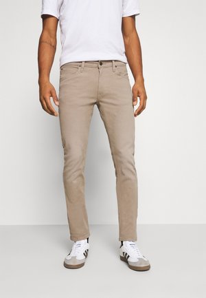 Homme portant un pantalon beige slim, un t-shirt blanc et des baskets blanches avec des rayures noires, debout devant un fond uni.