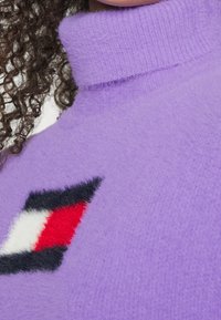 Cheveux bouclés à côté d'un pull à col roulé violet duveteux présentant un motif géométrique doux en noir, blanc et rouge sur la poitrine.
