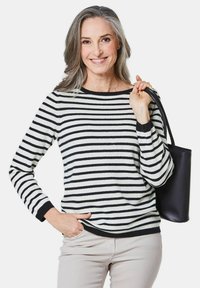 Schwarzer und weißer gestreifter Pullover mit langen Ärmeln und rundem Ausschnitt; eine schwarze Tasche wird in der linken Hand gehalten.