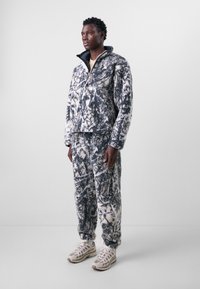 Fuzzy, black and white tie-dye patterned fleece jacket with a high collar, paired with matching pants and beige athletic shoes.