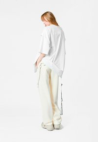 Camisa blanca oversized de corte holgado con un bolsillo trasero, combinada con pantalones crema que tienen un detalle de cadena y calzado gris claro.