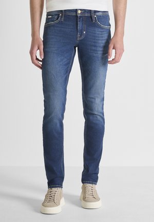 Vaqueros ajustados azules para hombre con un ligero desvanecimiento, combinados con zapatillas beige con cordones y una camiseta blanca, de pie sobre un fondo claro y sencillo.