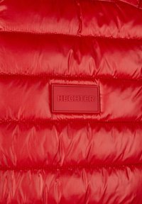 Rote Puffjacke mit gestepptem Design, ausgestattet mit einem rechteckigen gummierten Etikett mit der Aufschrift "HECHTER". Glattes, glänzendes Material mit sichtbaren Nähten.