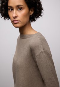 Hnědý pletený svetr s kulatým výstřihem, zdobený strukturovaným vroubkovaným lemem a pohodlným střihem. Měkká tkanina a volný střih.