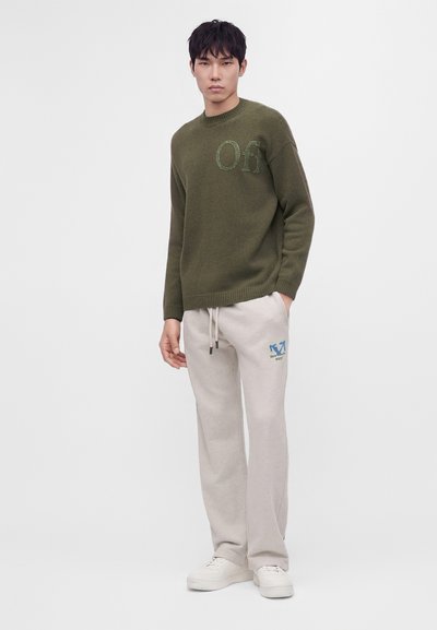 Jeune homme portant un pull vert olive avec les lettres "Off" et un pantalon de survêtement écru avec un motif bleu, debout devant un fond clair uni.