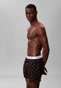 Homme musclé portant un boxer noir Calvin Klein avec une ceinture blanche, debout devant un fond gris uni, regardant vers la caméra.