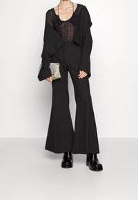 Kvinna i genomskinlig svart topp, oversized svart jacka, vida svarta byxor, svarta plattformstövlar, håller en grå clutch mot en vit bakgrund.