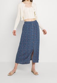 Sininen kukkakuvioinen maksihame, jossa on joustava vyötärönauha, edessä nappikiinnitys ja sivuhalkio; yhdistetty kermaiseen kuvioituun neuletakkiin ja sandaaleihin.