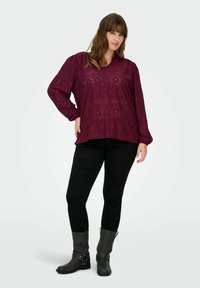 Burgundska dolga majica z dolgimi rokavi in vzorcem lukenj, sproščenega kroja, v kombinaciji s črnimi tesnimi kavbojkami in črnimi visokimi škornji z naramnicami.