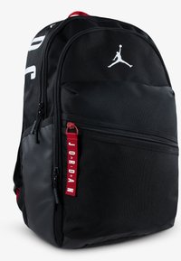 Mochila negra con una textura suave, que presenta un logo blanco de Jumpman, un tirador de cremallera rojo y un gran bolsillo frontal con acentos multicolores.