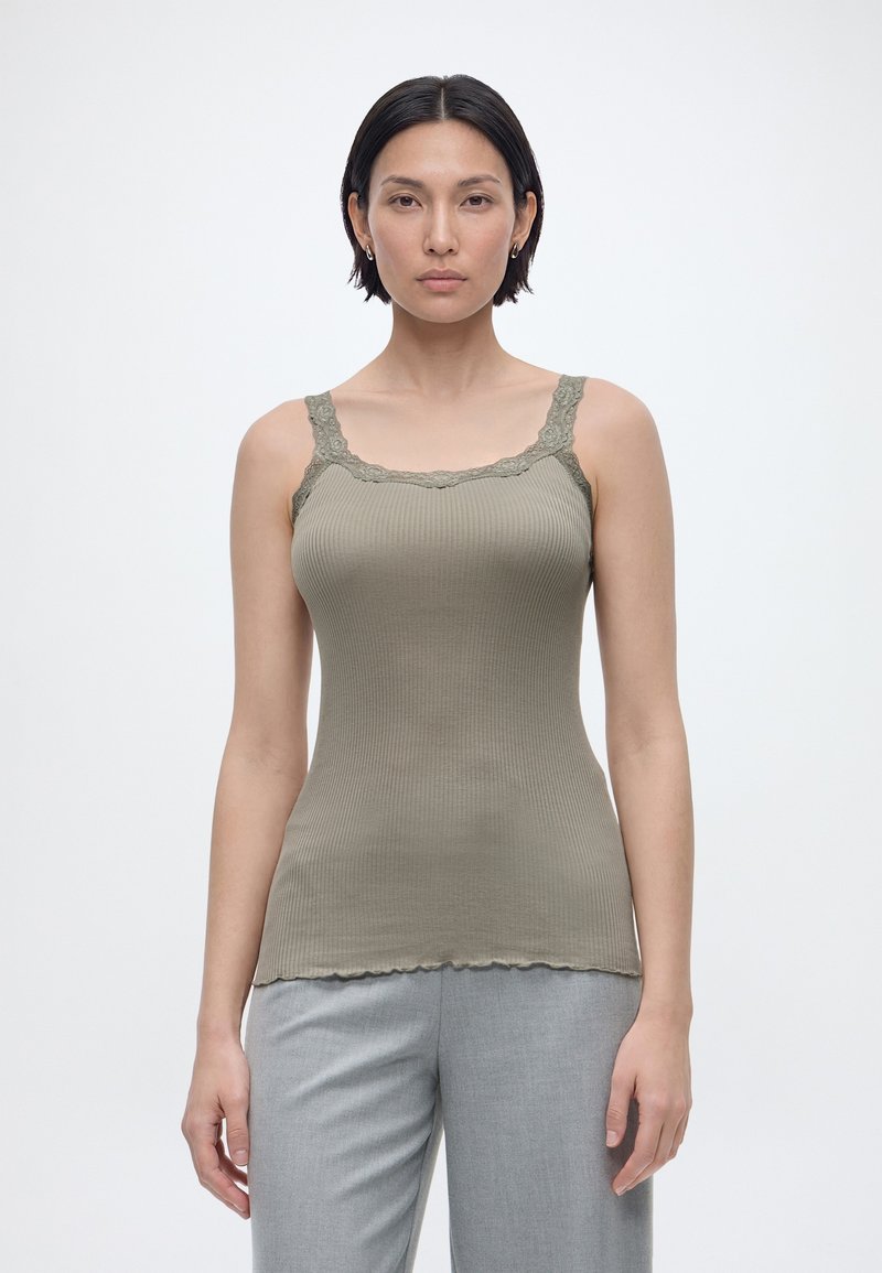 Γυναίκα με κοντά σκούρα μαλλιά φοράει ελαιί πράσινη ριμπ αμάνικη μπλούζα με δαντελωτή επένδυση και ανοιχτό γκρι παντελόνι, στέκεται μπροστά σε μονόχρωμο λευκό φόντο.