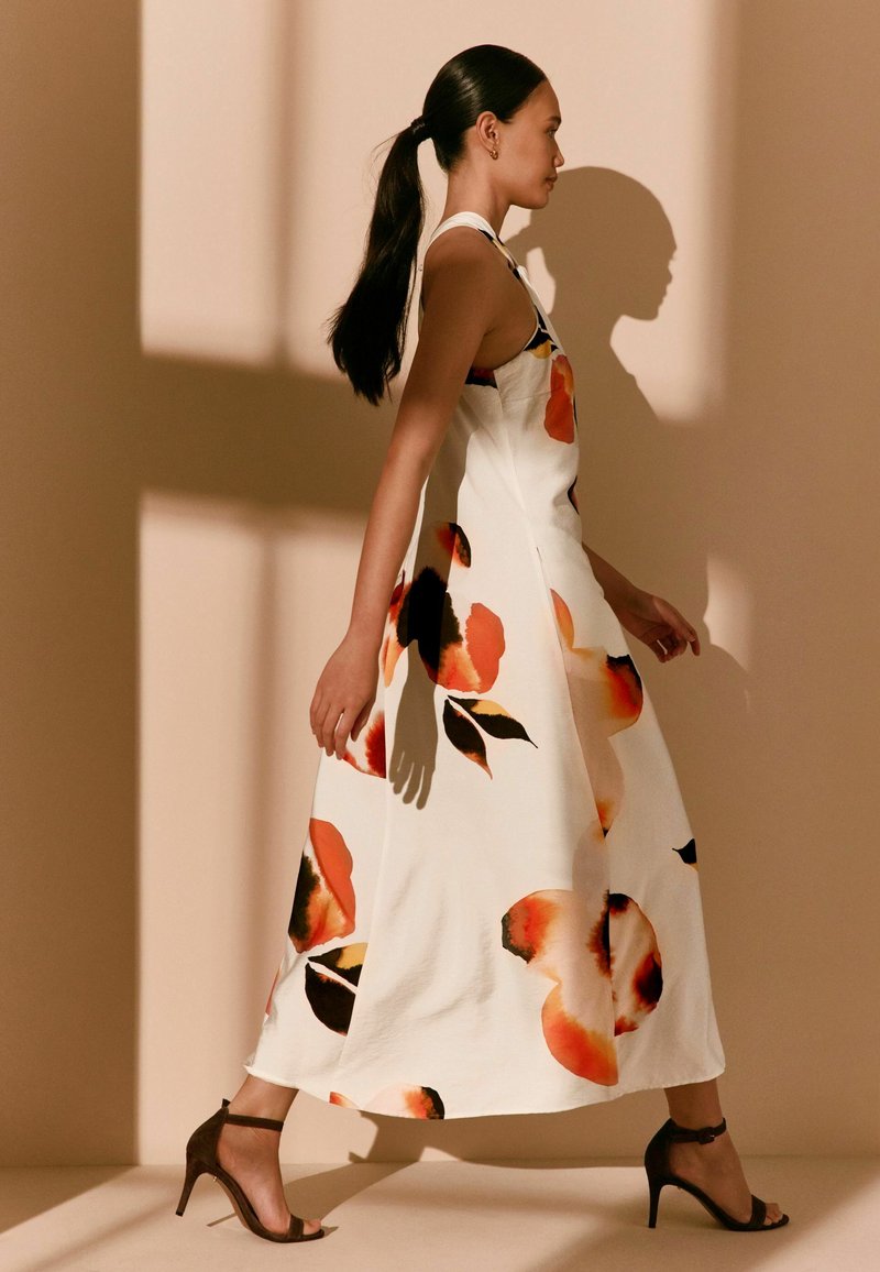Femme avec une longue queue de cheval foncée portant une robe blanche sans manches avec de grands motifs floraux orange et noirs et des sandales noires à talons, marchant.