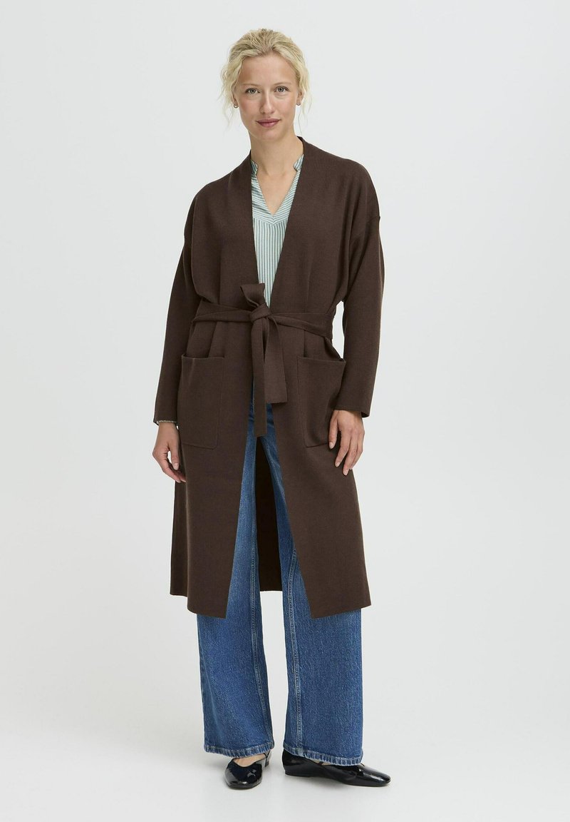 Donna che indossa un lungo cardigan marrone con cintura sopra una maglietta a strisce chiara, jeans larghi blu e scarpe piatte nere, in piedi su uno sfondo bianco.