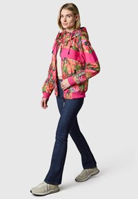 Woman walking wearing a colorful floral-patterned pink jacket, white crop top, dark blue jeans, and beige sneakers on a plain background.