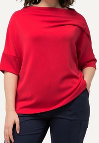 Röd off-the-shoulder-topp med draperad halsringning, mjuk textur och vida korta ärmar, i kombination med marinblå byxor med fickor.