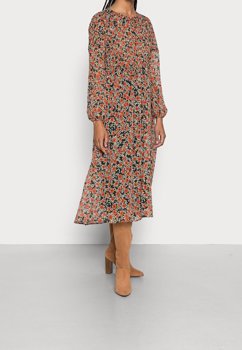 Vestido largo de manga larga en negro con flores rojas y blancas. Tela transparente, cintura fruncida y longitud a mitad de pantorrilla, combinado con botas de tacón alto en color beige.