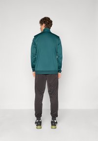 Treningjakke i mørkegrønn med hvite detaljer, lange ermer, høy krage, kombinert med svarte bukser og joggesko, glatt stoff, aktiv passform.