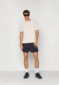 Lys beige kortermet skjorte, svarte shorts, hvite sokker og svarte treningssko. Modellen har på seg mørke solbriller og står foran en enkel bakgrunn.