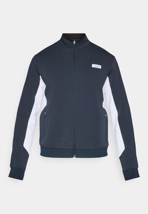 Marineblå zip-up jakke med hvide sidepaneler, lavet af blødt stof, med høj krave og to lynlåslommer.