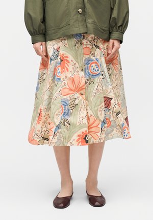 Jupe mi-longue avec imprimé floral en orange, bleu, vert et beige, portée avec une veste vert olive et des chaussures plates marron foncé.