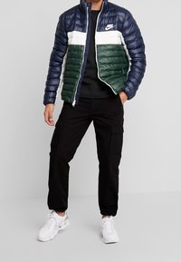 Vadderad Nike-jacka med färgblockering i marinblått, vitt och grönt. Har en dragkedja, hög krage och slät textur. Bärs över en svart skjorta och cargobyxor.