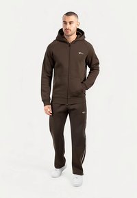 Brązowa bluza z zamkiem i kangurzą kieszenią z przodu, w zestawie z pasującymi spodniami z białymi paskami po bokach. Oba elementy wykonane z miękkiego, teksturowanego materiału.