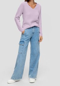 Pull en tricot lilas avec un col en V, associé à un jean cargo à jambes larges bleu clair avec poches latérales et coupe décontractée. Des baskets blanches complètent la tenue.