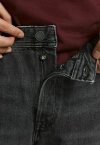 Mörkgrå denimjeans med knapp- och dragkedjestängning. Synlig sömdetaljering och lätt slitning vid midjan. Bärs med en vinröd topp.