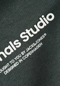 Čierna tkanina s textúrovaným vzorom, na ktorej je výrazne zobrazený biely text "JACK & JONES" a "Essentials Studio".