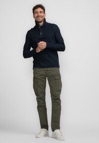 Marineblauer Strickpullover mit hohem Kragen und Knopf, kombiniert mit olivgrünen Cargo-Hosen mit Seitentaschen und weißen Sneakers.
