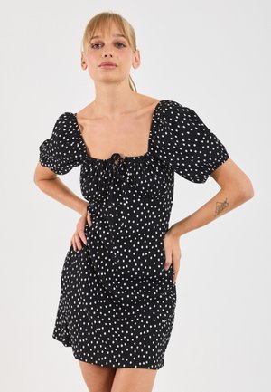 Femme aux cheveux blonds portant une robe noire à pois blancs et à manches bouffantes, se tenant les mains sur les hanches contre un fond blanc.