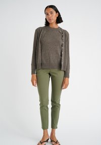 Cardigan marrone sopra un maglione marrone, abbinato a pantaloni verde oliva. Il cardigan ha un collo rotondo e bottoni sul davanti.