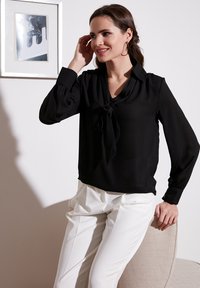 Black blouse with a bow tie detail at the neckline, long sleeves with button cuffs, paired with white tailored trousers, sitting on a chair.