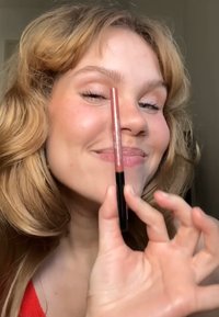 Crayon à lèvres dans un tube mince de couleur rose doux, avec un fond noir. Un modèle le tient près de sa joue, souriant.