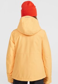 Gul vandtæt jakke med hætte, præget af et slankt design og udtalte sømme. Båret med en orange strikket hue.