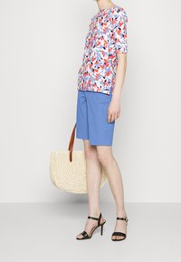 Kvinna som bär en blommig kortärmad topp, blå knälånga shorts, svarta högklackade sandaler och håller en vävd strandväska med bruna handtag.