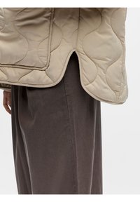 Μπεζ καπιτονέ μπουφάν με καμπυλωτές ραφές και μικρή σχισμή πάνω από σκούρο καφέ φαρδιά παντελόνια, που δείχνει μέρος ενός χεριού.