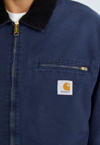 Mornaroplava jakna s crnim kabelastim ovratnikom, s zlatnim patentnim zatvaračem i prednjim džepom. Carhartt logotip na prsima.