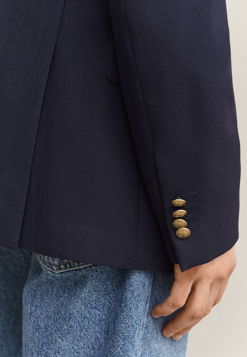 Marineblå blazerærme med fire gyldne dekorative knapper, båret over lyseblå denimjeans, hånd afslappet ved siden.