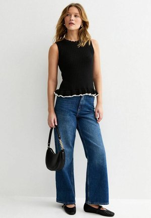 Kobieta ubrana w czarny dziergany top bez rękawów z falbaną na dole, niebieskie szerokie spodnie dżinsowe, czarne baleriny, trzymająca czarną torebkę.