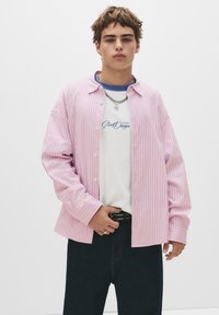 Cămașă butonată cu dungi roz pal, cu mâneci lungi, suprapusă pe un tricou alb. Pantaloni din denim închis și o curea neagră cu țepi. Colier din lanț argintiu.