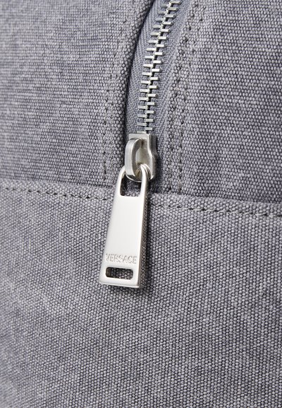 Saco têxtil cinza com puxador de zíper prateado com a gravação "VERSACE". O detalhe em primeiro plano mostra a textura suave e a costura robusta.