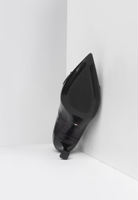 Black high-heel shoe viewed from below, featuring a pointed toe, textured sole, and small square heel. Smooth leather material.