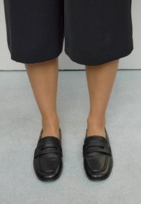 Person wearing black leather slip-on shoes and black cropped pants standing on a gray floor.